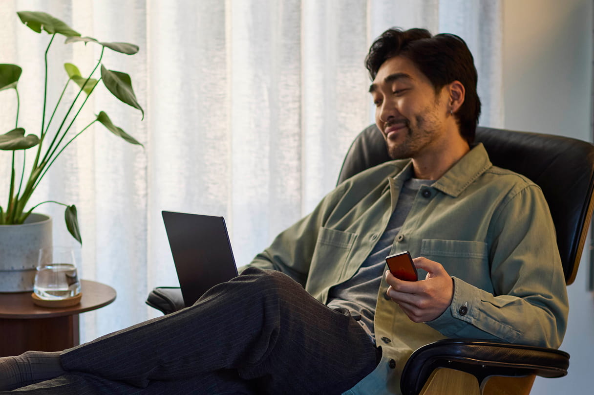 男の人が椅子に座り、MacBookを使いながら、片手に Trezor Safe 7 のハードウェアウォレット を持っています。