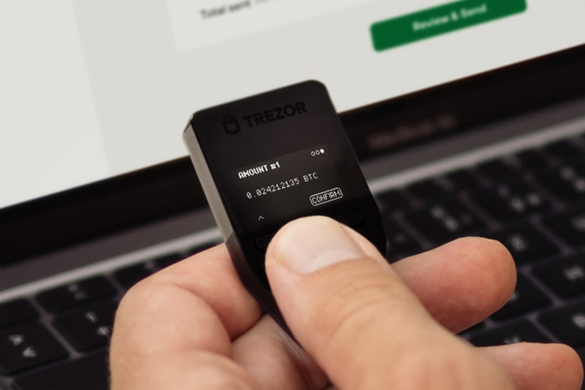 ノートパソコンの画面の前で、ビットコインの取引金額を表示している Trezor Safe 3 のハードウェア暗号資産ウォレットを手に持っている様子。