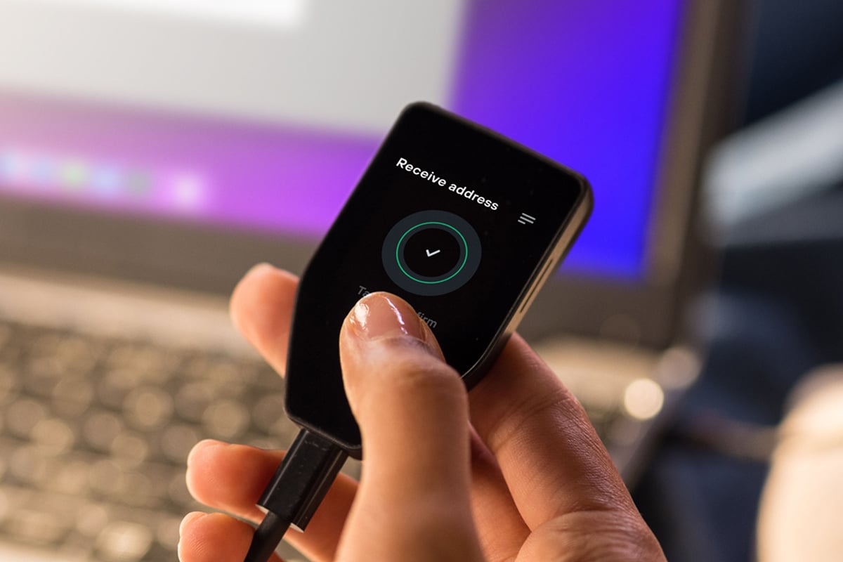 USBで接続されたTrezor Safe 5ハードウェアウォレットを手に持ち、「受取アドレス」の確認画面にチェックマークが表示されている様子のクローズアップ。背景にはぼかしたノートパソコンが映っている。