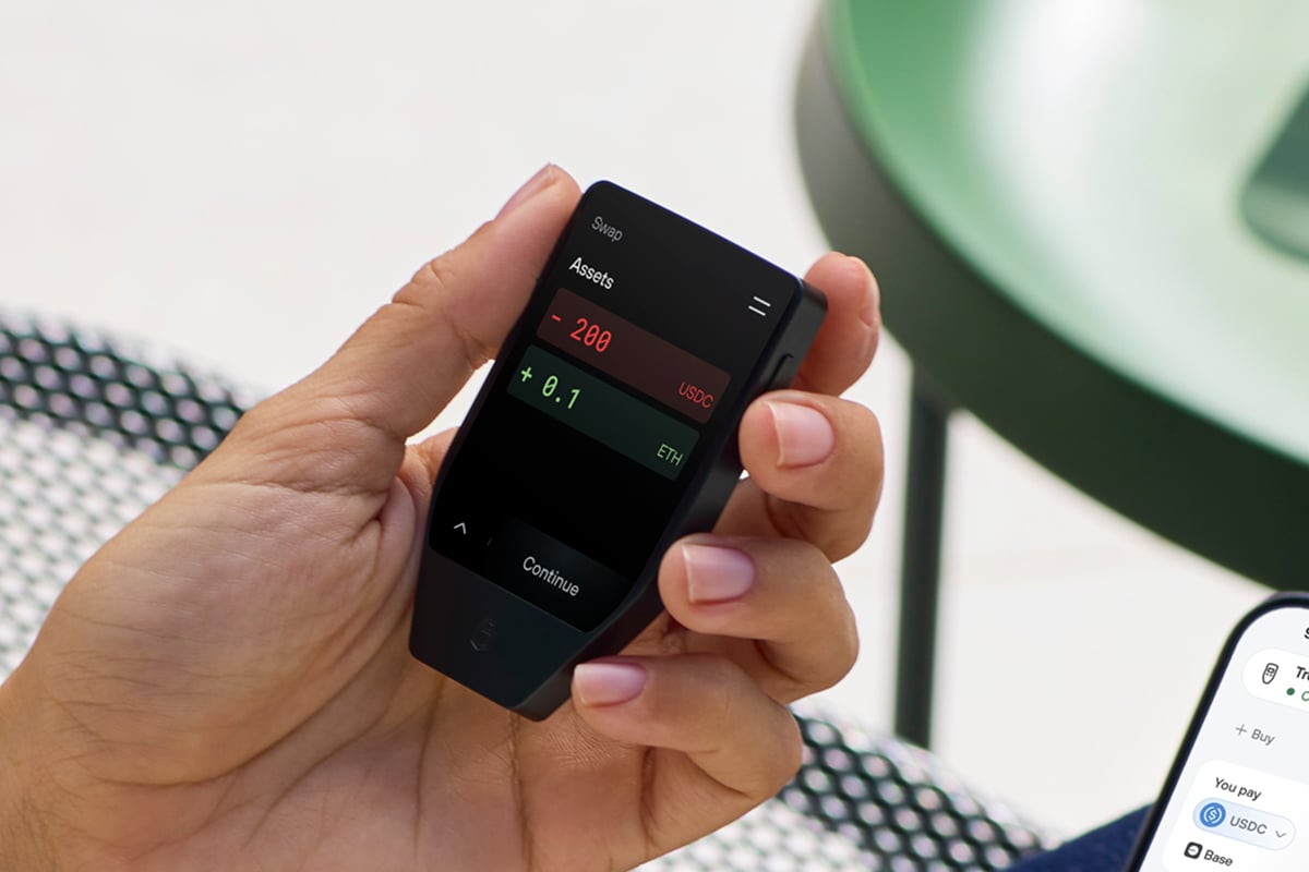 手に持った Trezor Safe 7 ハードウェアウォレットに、USDC と ETH の残高が表示された暗号資産のスワップ画面が映し出されており、柔らかな室内の背景を背にしている。