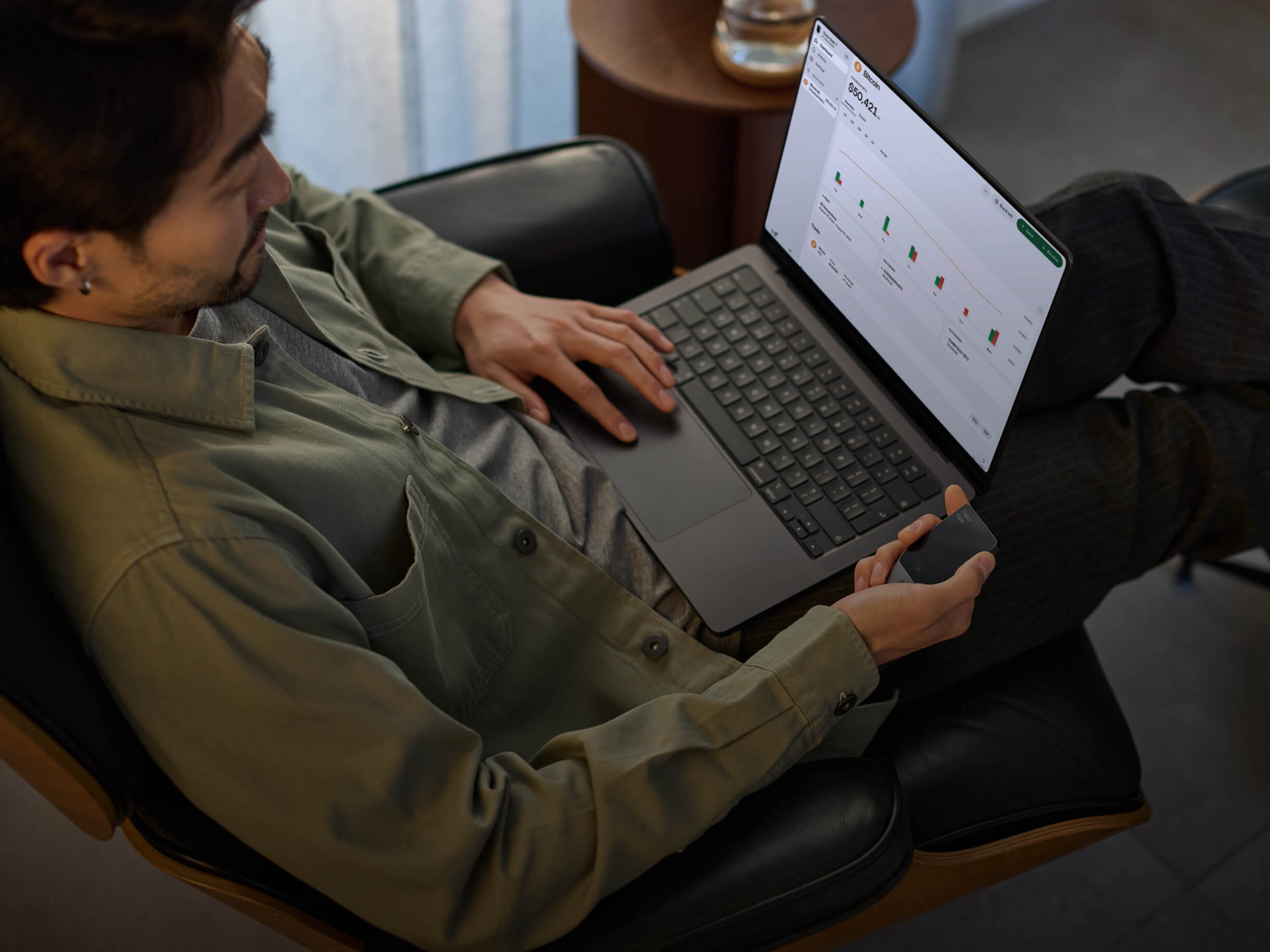A man sits on a chair using a MacBook while holding a Trezor Safe 7 hardware wallet in one hand.
