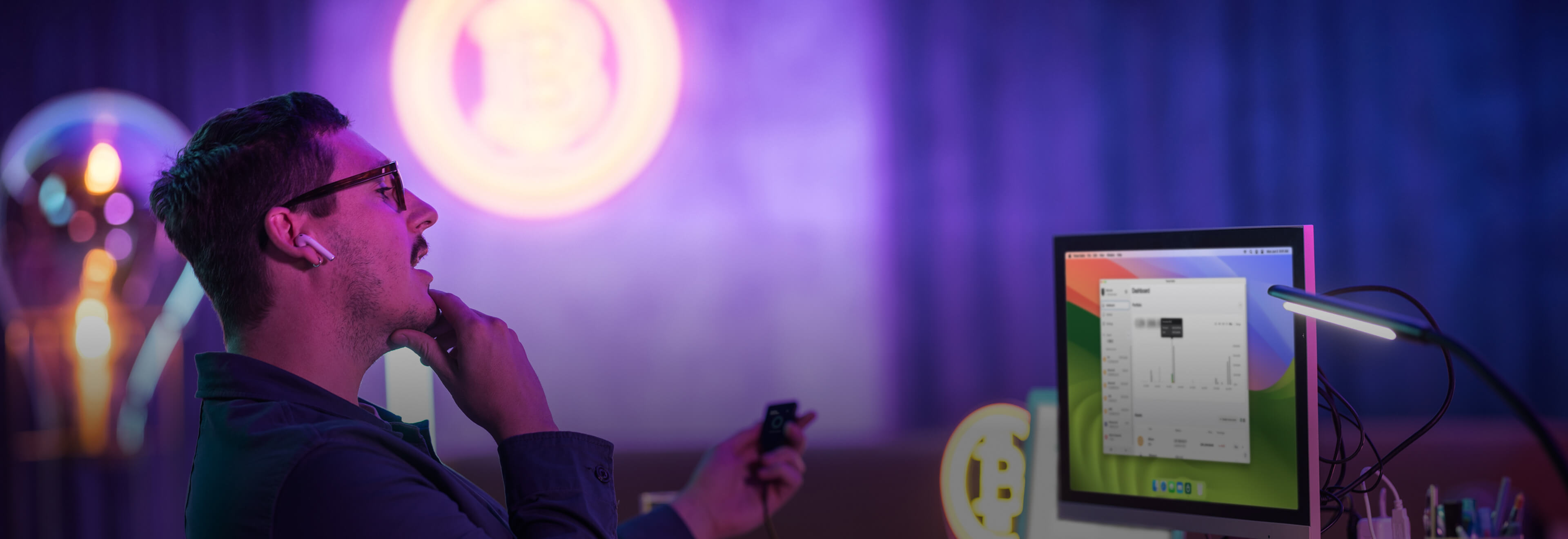 A man wearing glasses and wireless earbuds sits at a desk, thoughtfully looking at a computer monitor displaying a Trezor Suite dashboard. He holds a Trezor Save 5 in one hand and rests his chin on the other. The background features neon Bitcoin symbols and a dimly lit room with purple and blue lighting, creating a modern, tech-focused atmosphere.