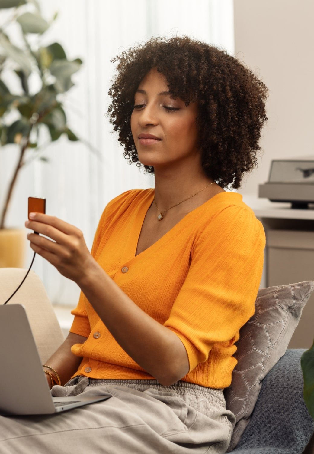 Woman sitting on a sofa using a laptop and holding a Trezor Safe 5 hardware wallet for secure cryptocurrency storage at home, with plants and modern decor in the background.