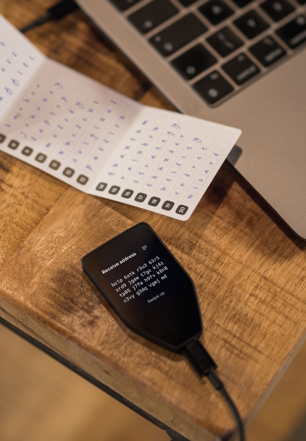 Trezor Safe 5 hardware wallet connected to a laptop via USB cable, placed on a wooden desk next to a recovery seed card with handwritten seed words.