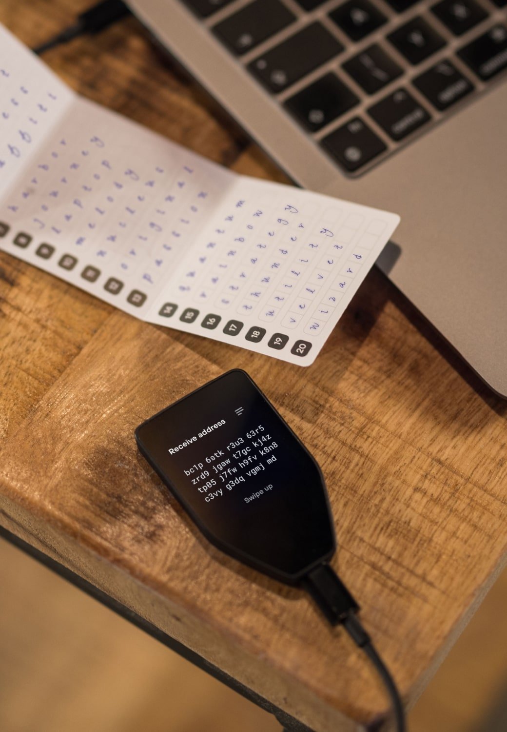 Trezor Safe 5 hardware wallet connected to a laptop via USB cable, placed on a wooden desk next to a recovery seed card with handwritten seed words.