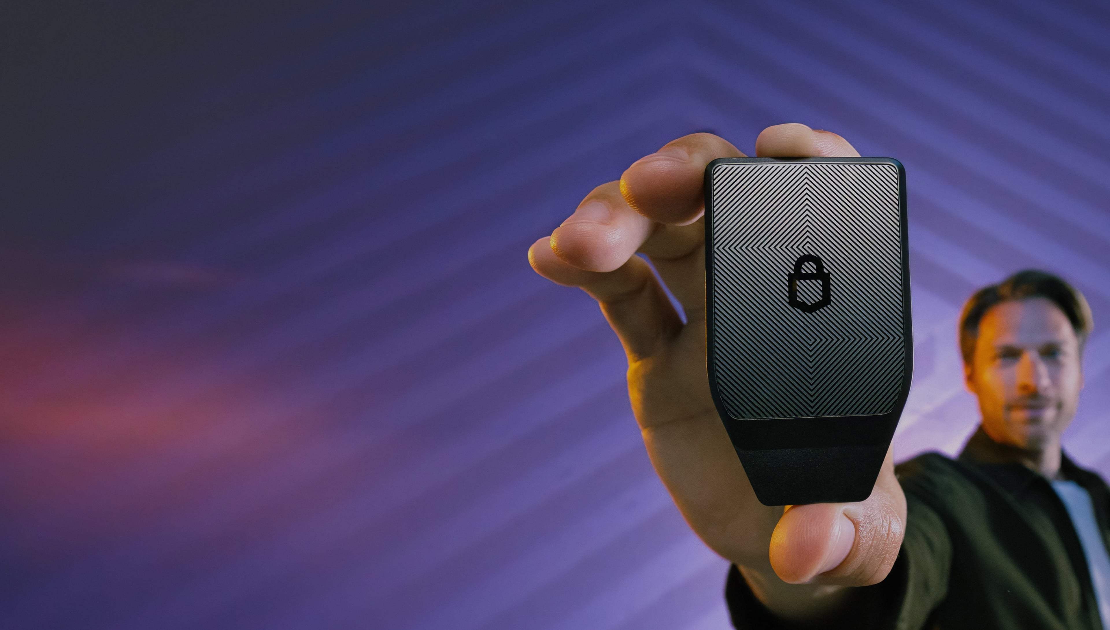 A hand holding a Trezor Safe 7 hardware wallet against a purple patterned background.