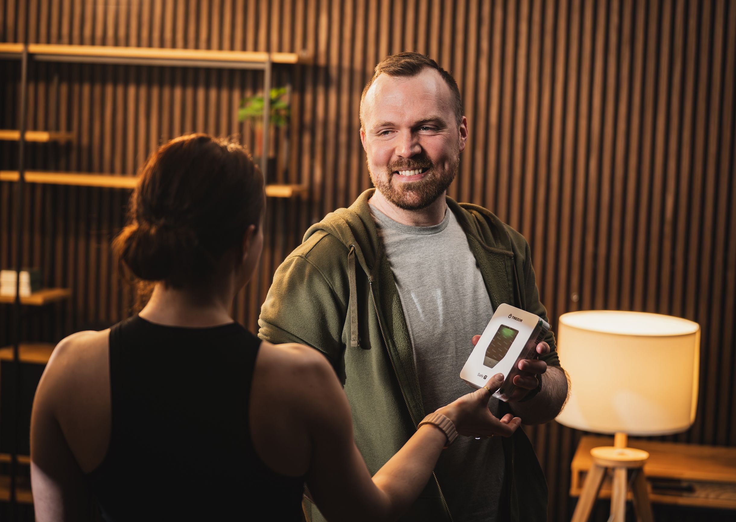 A man in a green hoodie smiling and handing a Trezor Safe 7 package to a woman in a modern room with slatted wood walls.